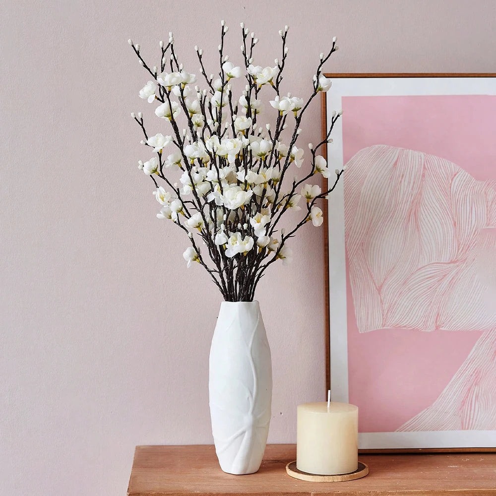 A vase filled with white artificial plum blossoms sits on a wooden surface next to a pink abstract framed print.