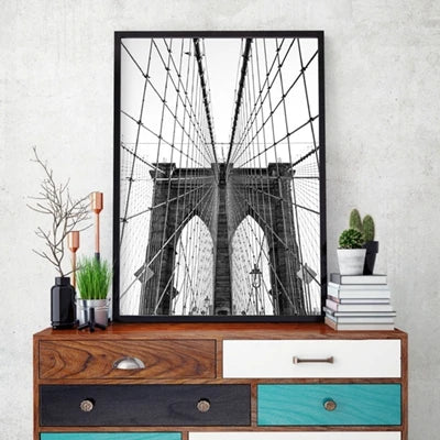 Black and white photograph of the Brooklyn Bridge framed in a black canvas print displayed on a wooden cabinet.