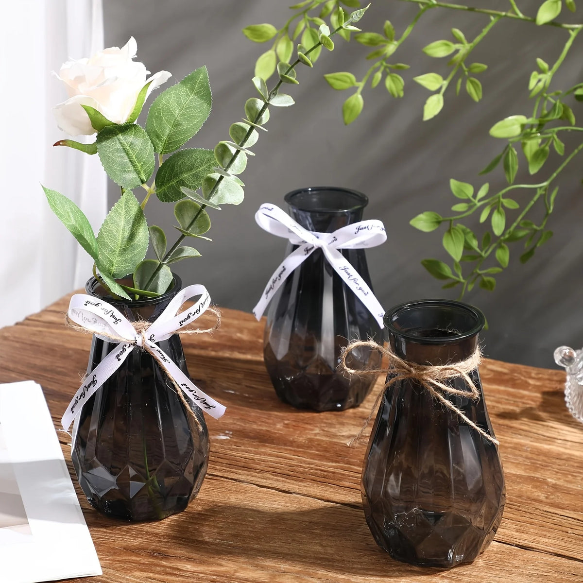 Three faceted diamond crystal vases with natural greenery and white ribbon on a wooden table.