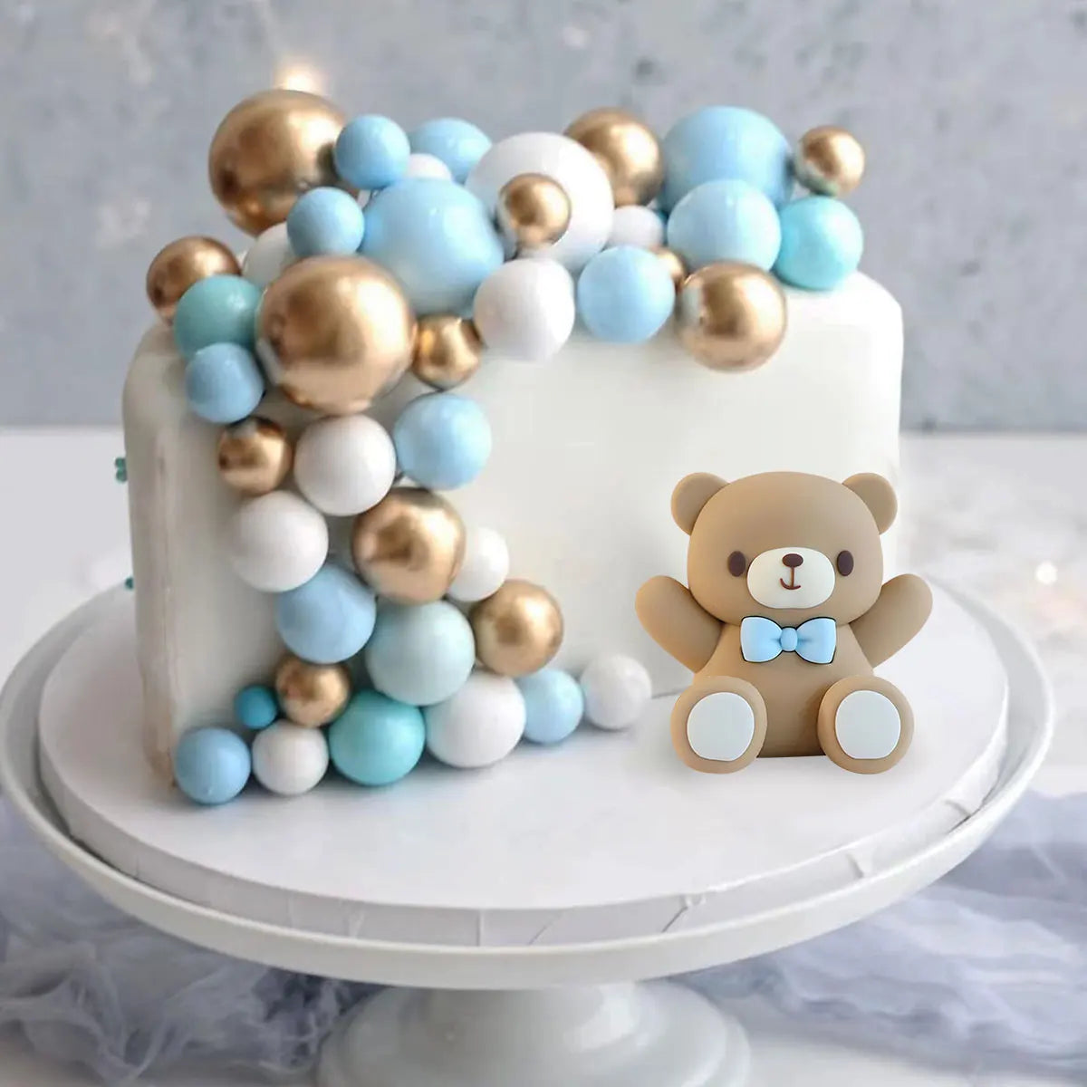 A white cake decorated with round blue and gold edible pearl candies and a small bear figure.