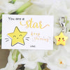 A card with the text You are a star keep shining and a yellow star keychain hanging from a silver ring surrounded by white fl