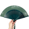 Elegant bamboo folding fan in dark green with a classic design being held by a hand.