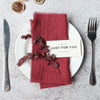 A set of ten handcrafted fringed cotton napkins in a deep reddish-pink color placed on a white plate with cutlery.