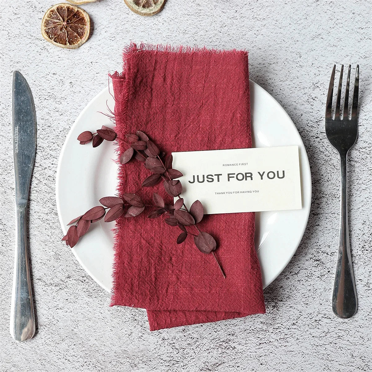 A set of ten handcrafted fringed cotton napkins in a deep reddish-pink color placed on a white plate with cutlery.