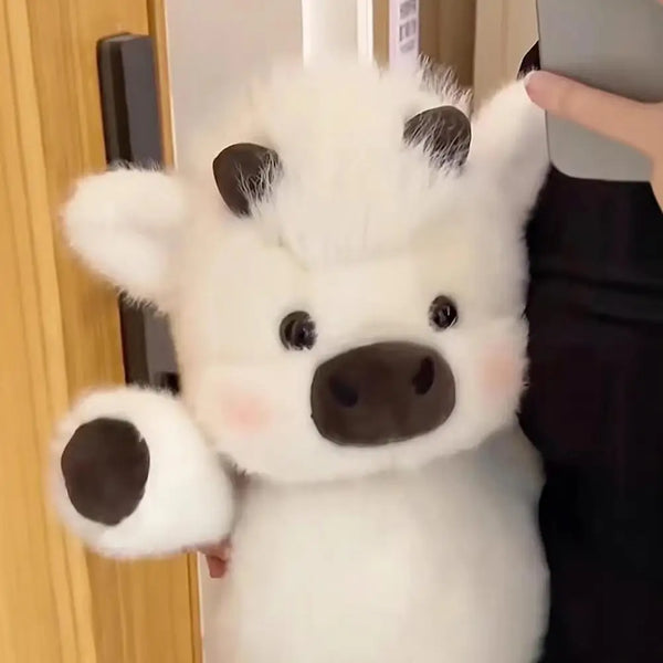 A white plush toy shaped like a pig or calf with dark ears and a snout is being held by a hand.