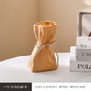 A small, light brown, fabric money bag shaped vase is displayed on a white surface.