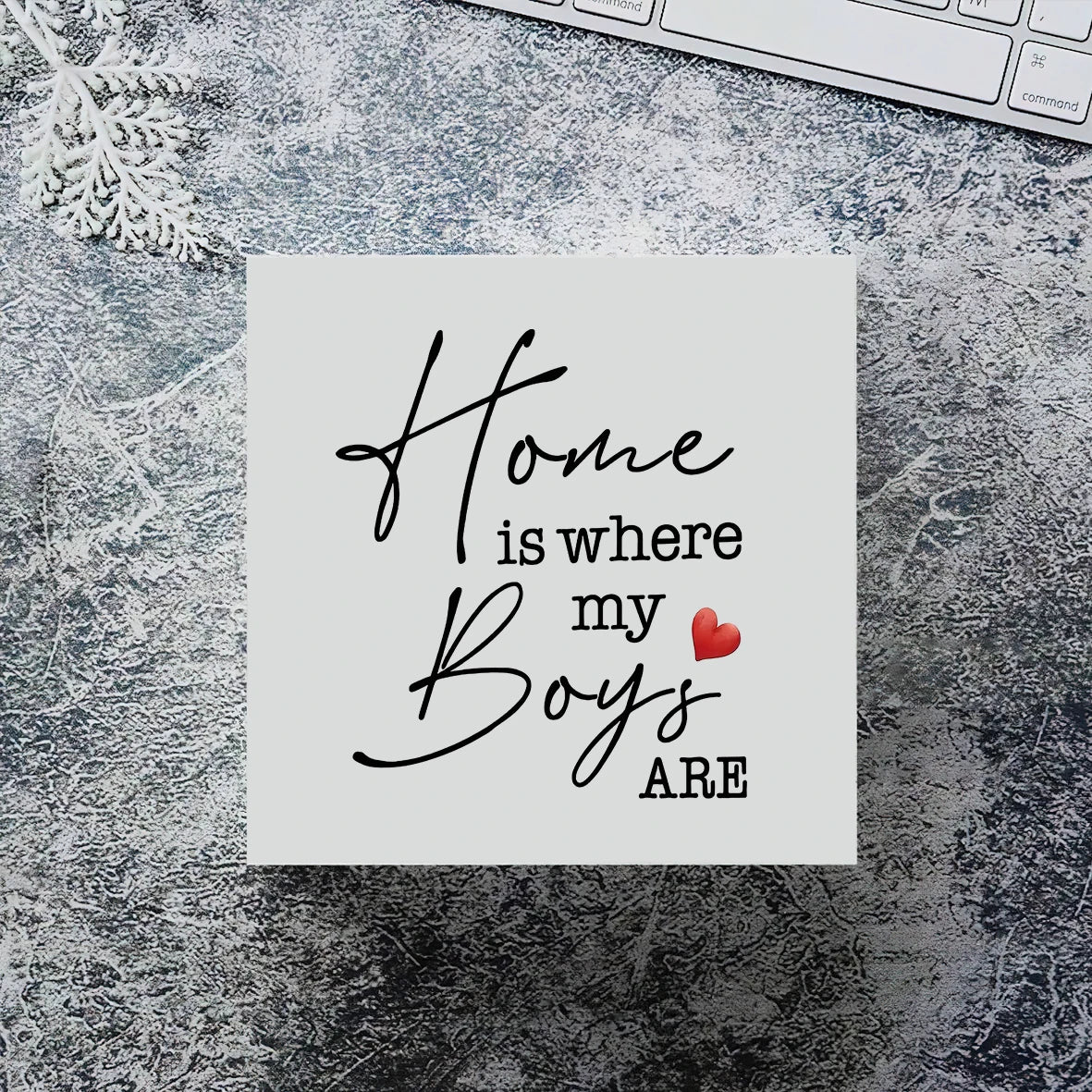 A white sign with elegant script reads Home is where my boys are with a small red heart on a textured surface.