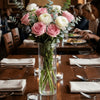 A clear glass cylinder vase filled with green stems and foliage on a wooden table.