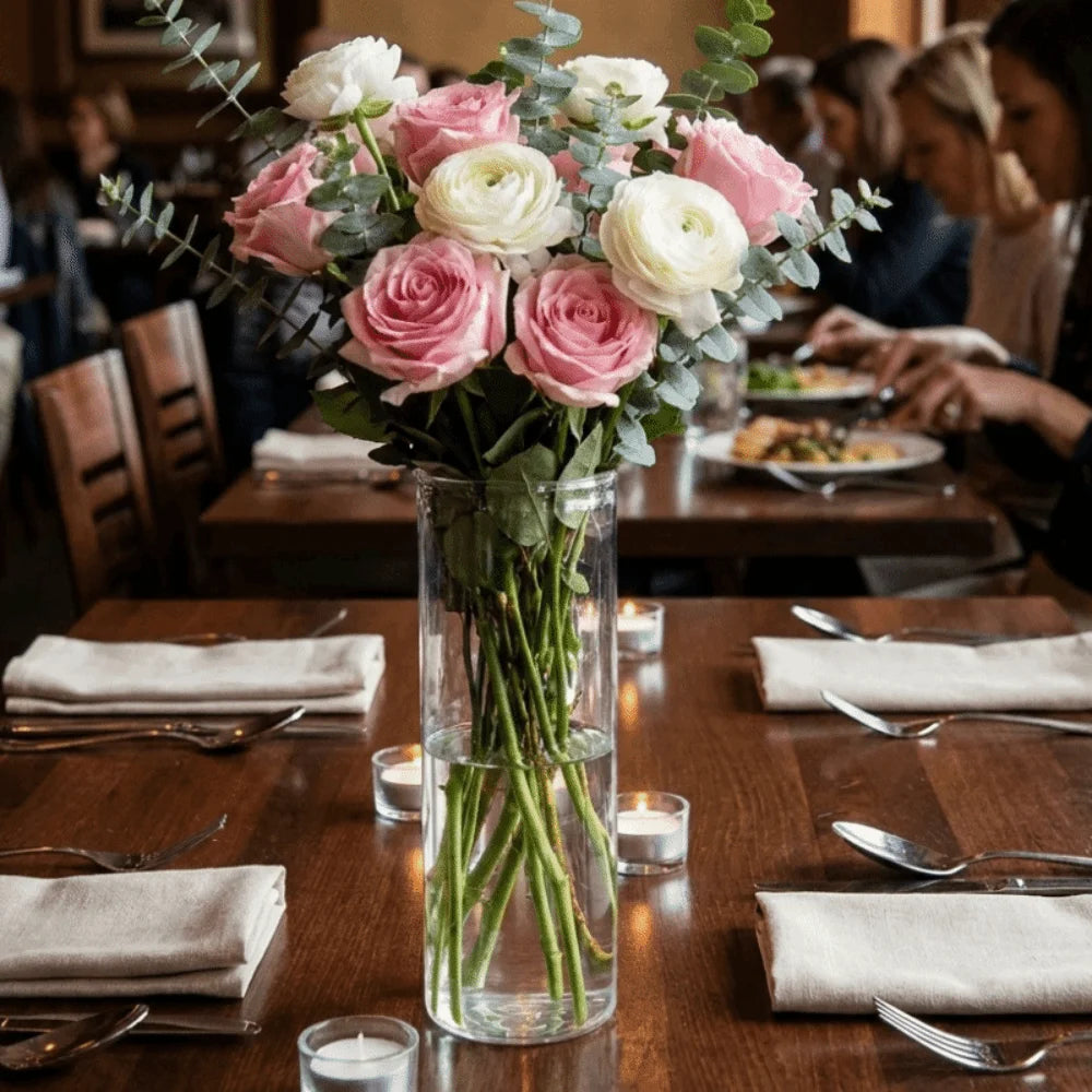 A clear glass cylinder vase filled with green stems and foliage on a wooden table.
