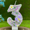 A glittery purple number three candle decorated with small flowers and a large purple butterfly on a white base.