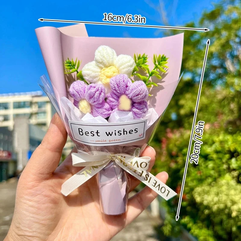 A hand holds a bouquet of handmade crochet flowers in pink paper with a white ribbon and a tag that reads Best wishes.