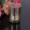 A clear glass votive candle holder with vertical fluted texture sits on a dark reflective surface next to pink and peach flow