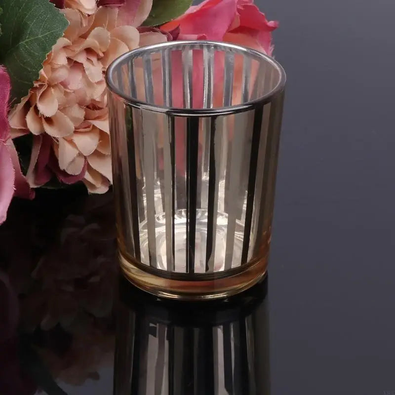 A clear glass votive candle holder with vertical fluted texture sits on a dark reflective surface next to pink and peach flow