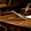 A vintage brass hummingbird statue rests on a wooden desk next to a feather quill and old books.