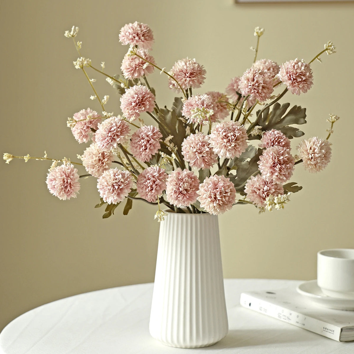 A bouquet of pink silk chrysanthemum artificial flowers in a white vase on a light surface.