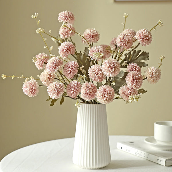 A bouquet of pink silk chrysanthemum artificial flowers in a white vase on a light surface.