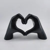 A black resin sculpture shaped like two hands forming a heart, displayed against a light gray background.