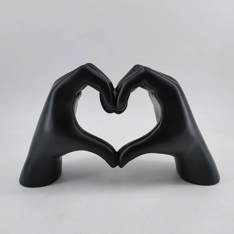 A black resin sculpture shaped like two hands forming a heart, displayed against a light gray background.