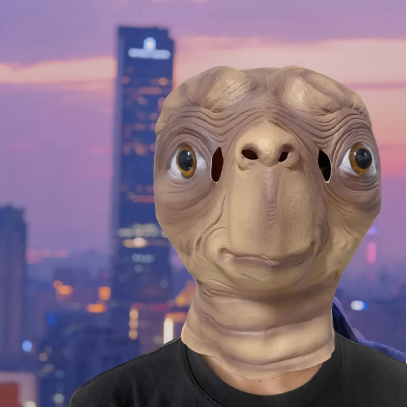 A realistic cute alien full-face latex mask with large eyes and wrinkled skin against a city skyline background.