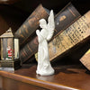 A white figurine of a praying angel with feathered wings is displayed on a wooden shelf among old books and a decorative lant