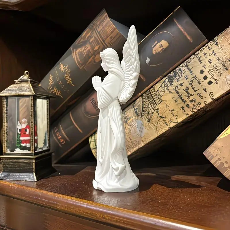 A white figurine of a praying angel with feathered wings is displayed on a wooden shelf among old books and a decorative lant