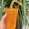 A hand holds an orange reusable sipper cup with a straw and lid against a background of green foliage.