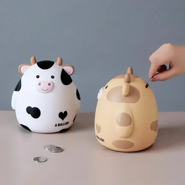 A white and black cartoon cow piggy bank with a gold-colored elephant-shaped coin holder next to some coins.