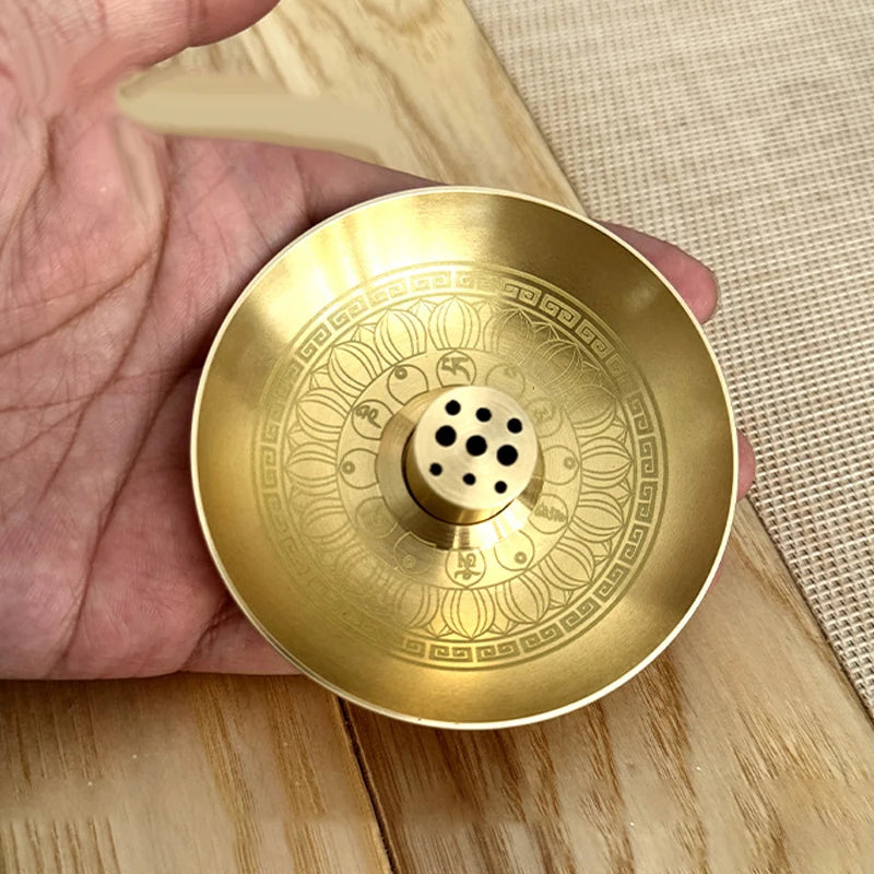A round brass incense stick holder plate with a decorative pattern is held in a hand.