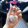 A hand holds a gold-colored keychain charm shaped like a sparkling pink heart.