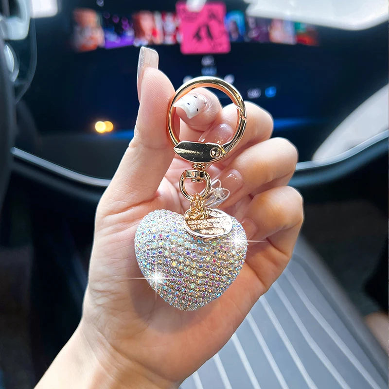 A hand holds a gold-colored keychain charm shaped like a sparkling pink heart.