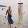 A woman walks along a shoreline while a hanging wind chime made of aluminum tubes and gold bells is visible.