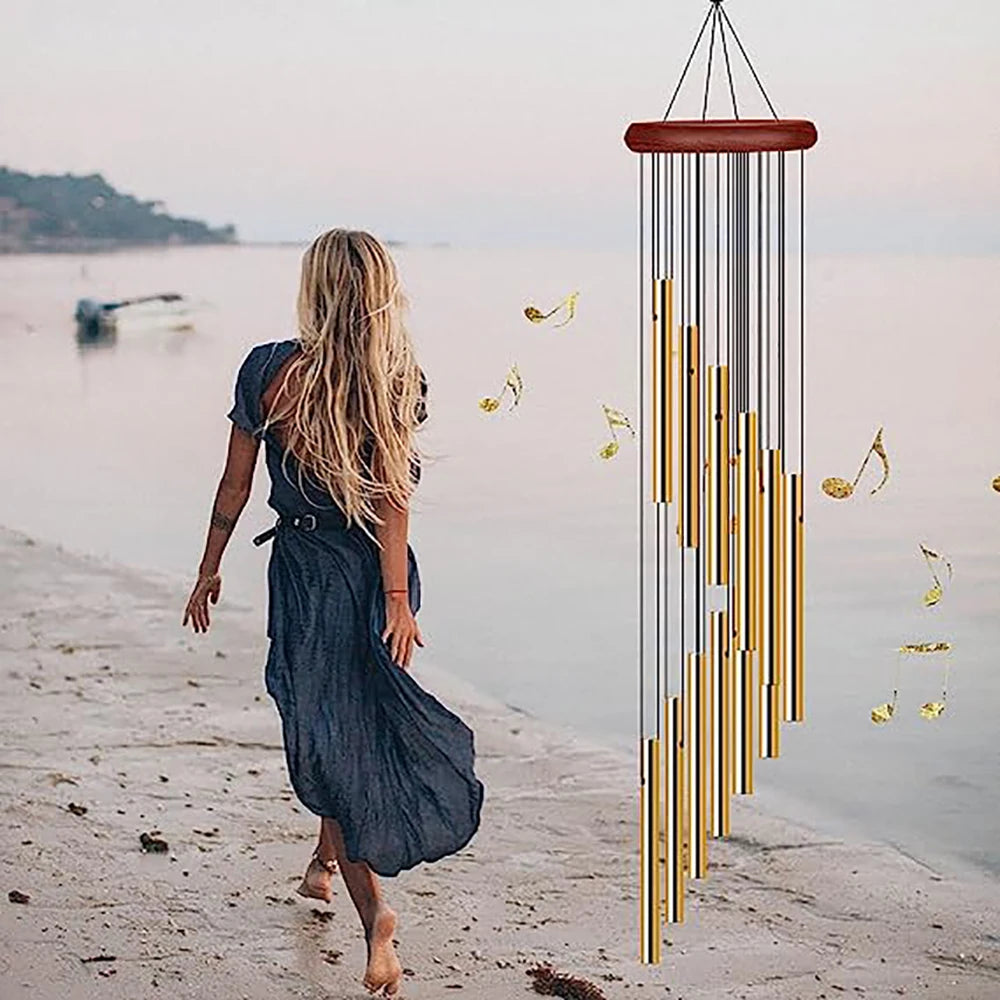 A woman walks along a shoreline while a hanging wind chime made of aluminum tubes and gold bells is visible.
