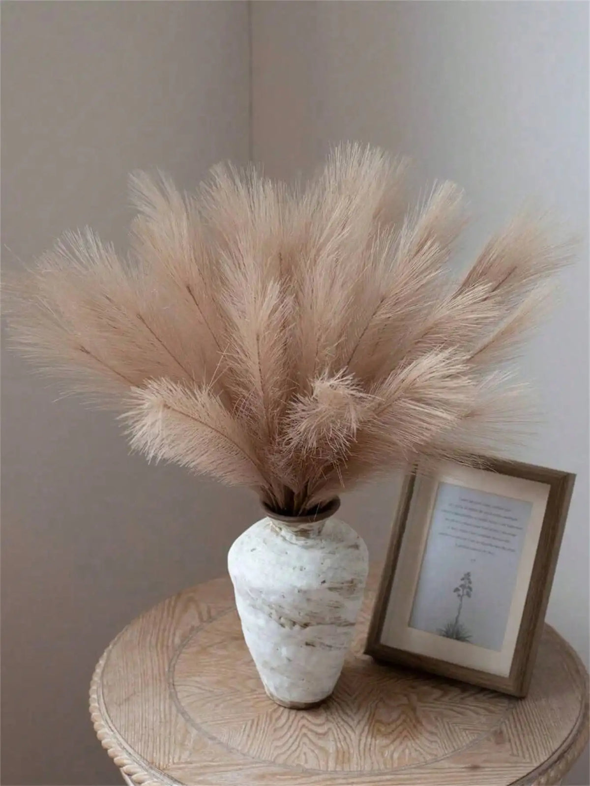 A bouquet of light beige pampas grass stems in a white ceramic vase on a wooden surface.