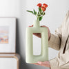 A person holds a pale green, geometric ceramic vase with a rectangular opening, displaying it against a light background.