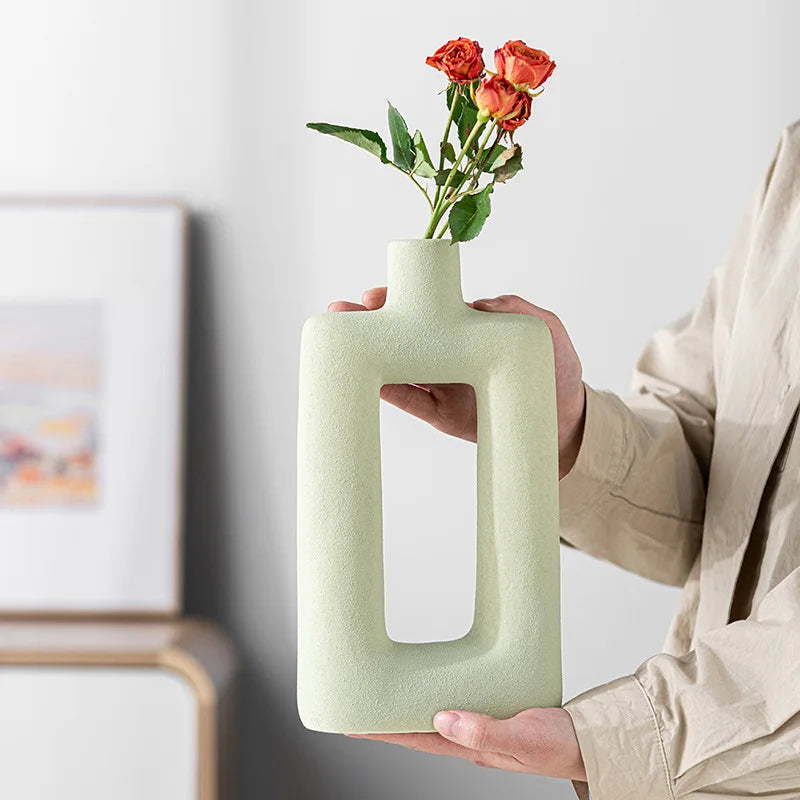 A person holds a pale green, geometric ceramic vase with a rectangular opening, displaying it against a light background.