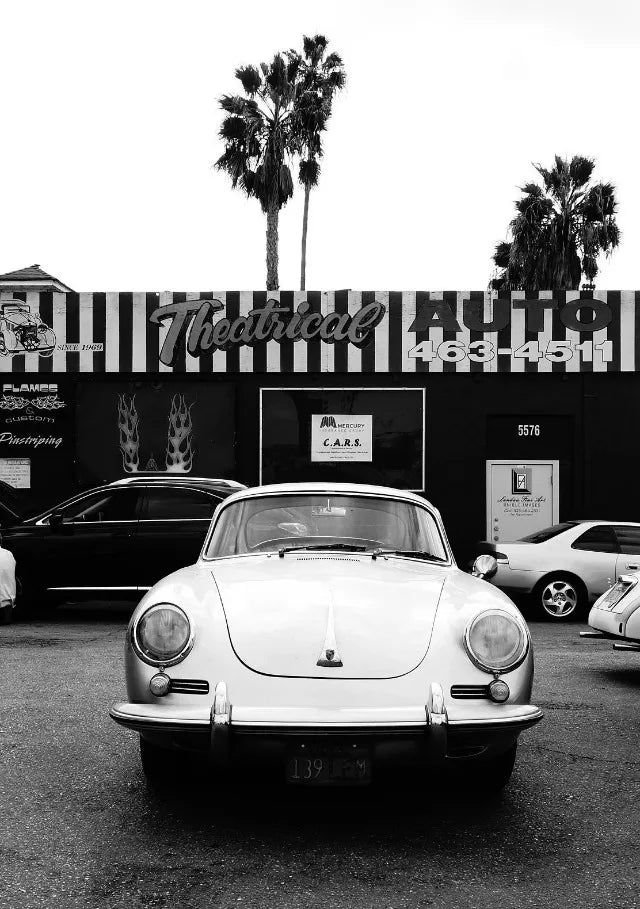 Une voiture blanche vintage est garée devant un bâtiment avec des palmiers et une enseigne.