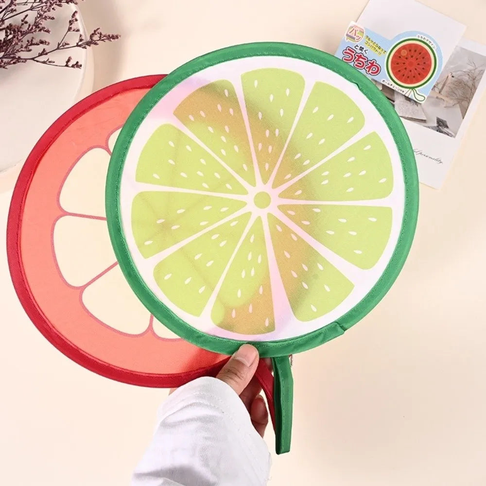 A round foldable hand fan with a green frame and a sliced citrus fruit design, held by a hand.