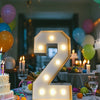 A large illuminated marquee number 2 made of LED lights sits on a table surrounded by balloons and candles.