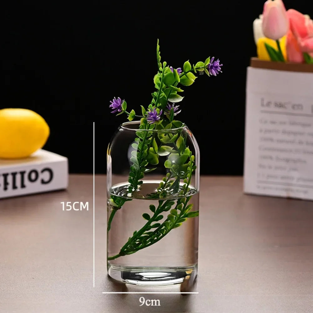 A clear glass hydroponic vase holds green foliage with purple flowers on a table.