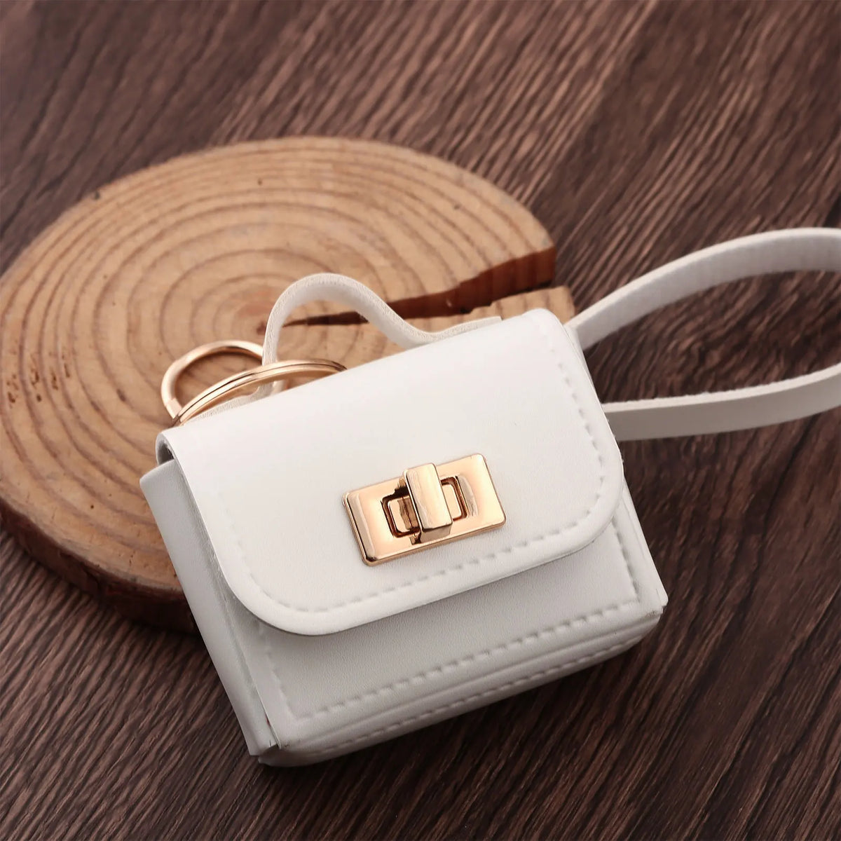 A small white leather openable bag with a gold-toned clasp rests on a dark wooden surface next to a gold ring.