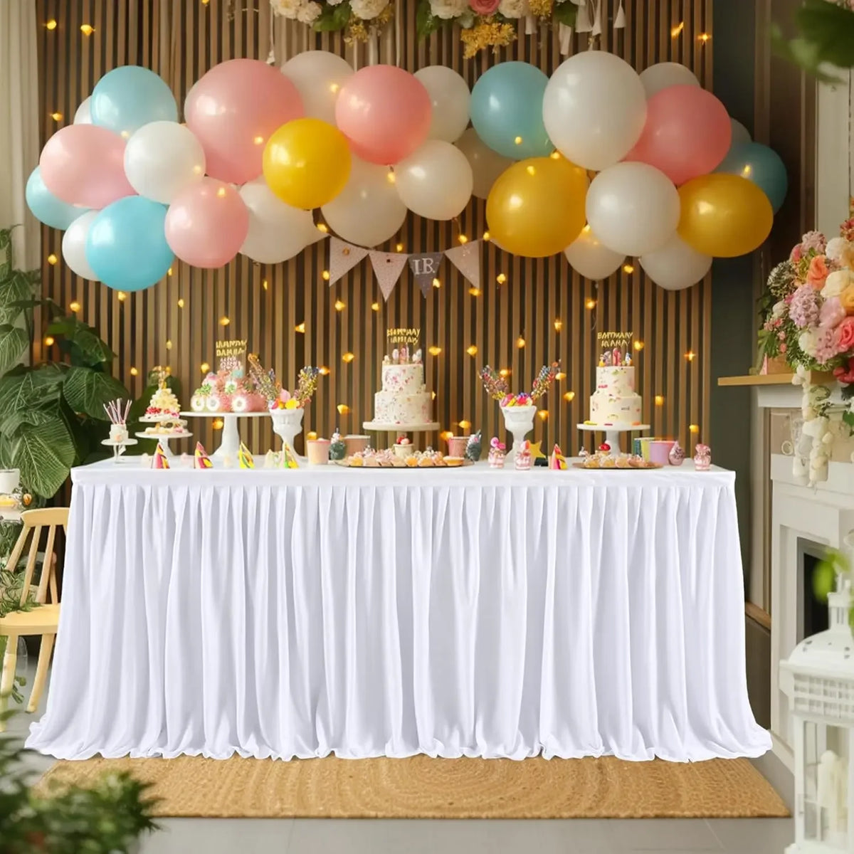A white pleated polyester table skirt decorated with pink and yellow balloons and cake toppers for a celebration.
