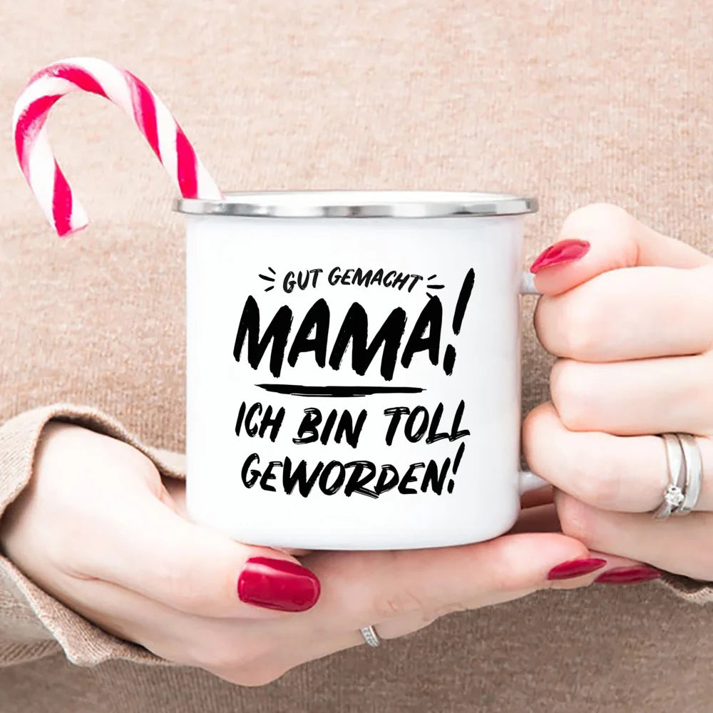 A white enamel mug with black text on it is held by a person, featuring the phrase Gut gemacht Mama ich bin toll geworden