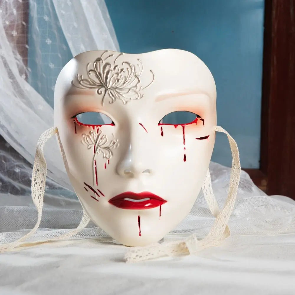 A white masquerade mask with intricate silver floral designs and red paint drips, displayed on a white cloth.