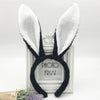A pair of black bunny ear headbands with white fluffy ears displayed in a white photo frame.