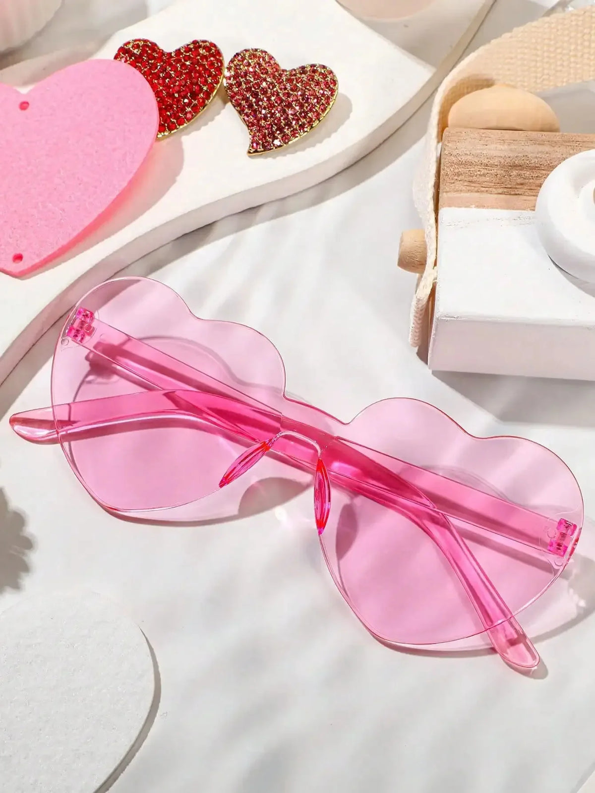 Pink rimless heart-shaped sunglasses resting on a white surface next to red hearts.