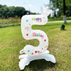A white foil balloon shaped like the number 5 with a crown and birthday text standing on green grass.