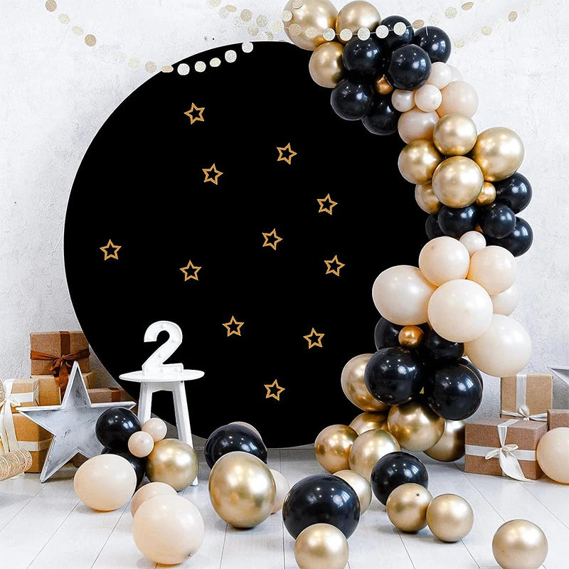 A dark circular photo backdrop decorated with gold stars and clusters of light beige, black, and gold balloons.
