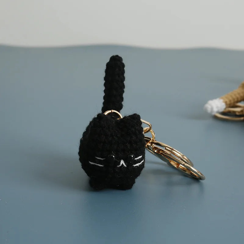 A black crocheted cat keychain pendant with a long tail and gold-toned hardware resting on a gray surface.