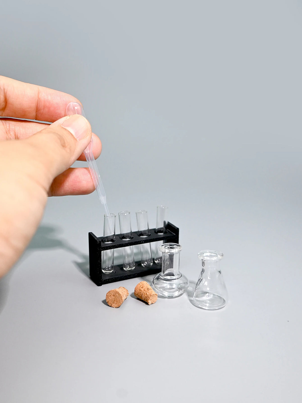 A hand is pipetting liquid into a set of miniature laboratory glassware for dollhouse or photography.
