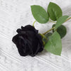 A single, dark black artificial rose with green leaves on a white textured background.